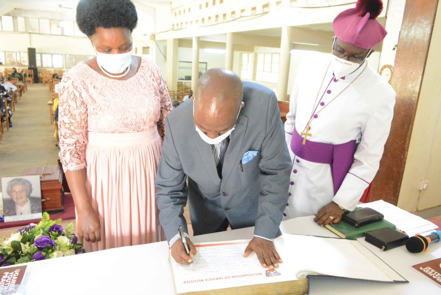 Mr. Jackson Katongore Bells escorted by the wife, signing Bishop Stuart University Recognition Service Book